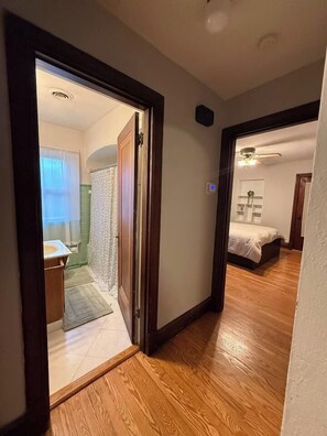 A hallway leading to a bedroom with a bed, a bathroom with a shower, and a wooden floor. - Historic STL Home near Wash U/Massive Backyard (University City)