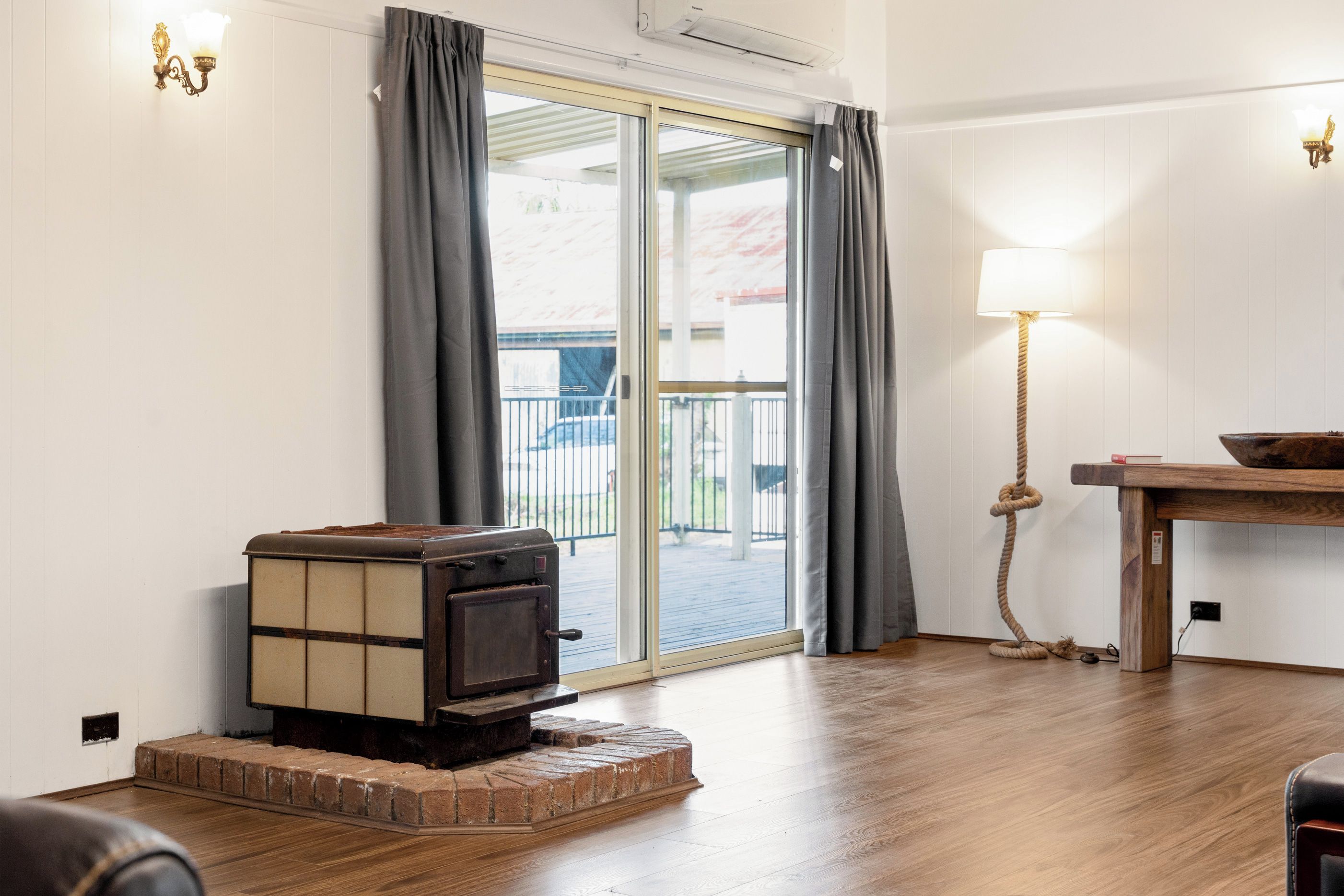 A room with a wood-burning stove, a sliding glass door to an outdoor area, a table with a lamp, and a brick base under the stove.