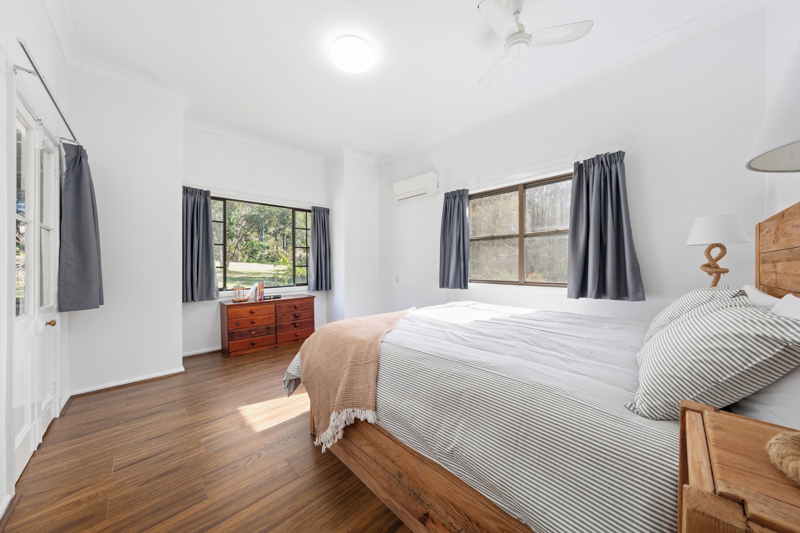 Un dormitorio con una cama de madera, una mesita de noche, un aparador y dos ventanas con cortinas.