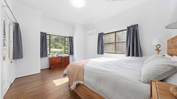 Un dormitorio con una cama de madera, una mesita de noche, un aparador y dos ventanas con cortinas.