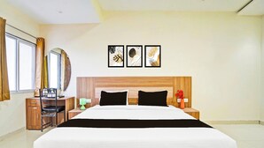 Ein modernes Hotelzimmer mit einem großen Bett, einem Holzkopfteil, einem Schreibtisch mit Stuhl, einem Nachttisch mit Lampe und einem Fenster mit Vorhängen.