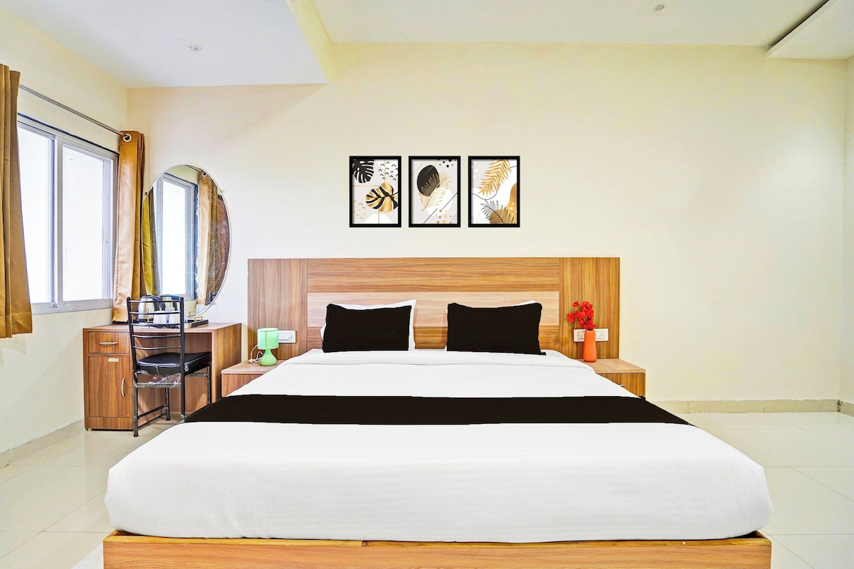 Ein modernes Hotelzimmer mit einem großen Bett, einem Holzkopfteil, einem Schreibtisch mit Stuhl, einem Nachttisch mit Lampe und einem Fenster mit Vorhängen.