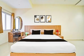 A modern hotel room with a large bed, a wooden headboard, a desk with a chair, a nightstand with a lamp, and a window with curtains. - Super Hotel Kalpi Formerl Gajanand (Gwalior)