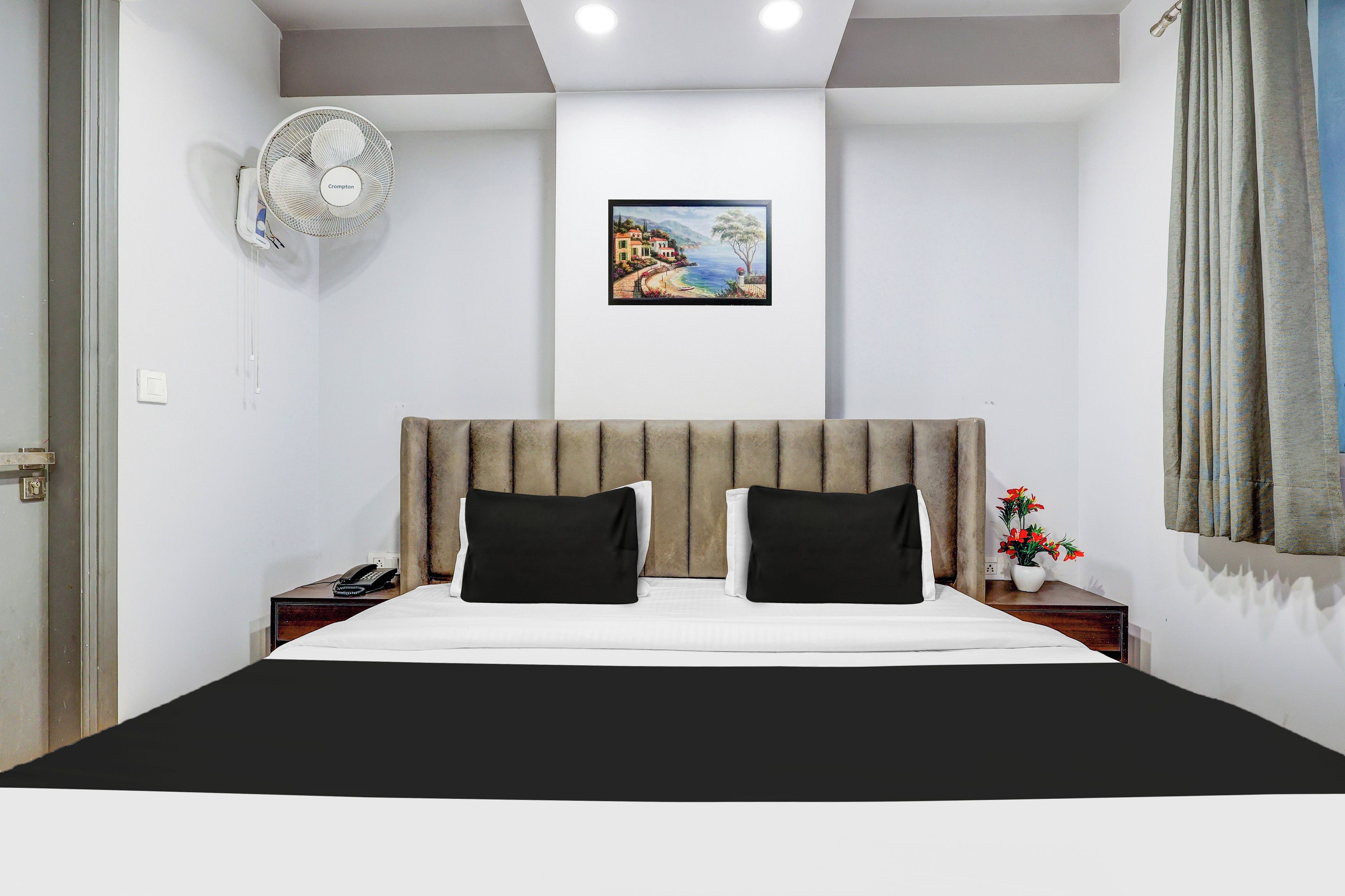 Une chambre d’hôtel avec un lit, une table de chevet, un téléphone, un ventilateur fixé au mur, un tableau et une fenêtre avec des rideaux.