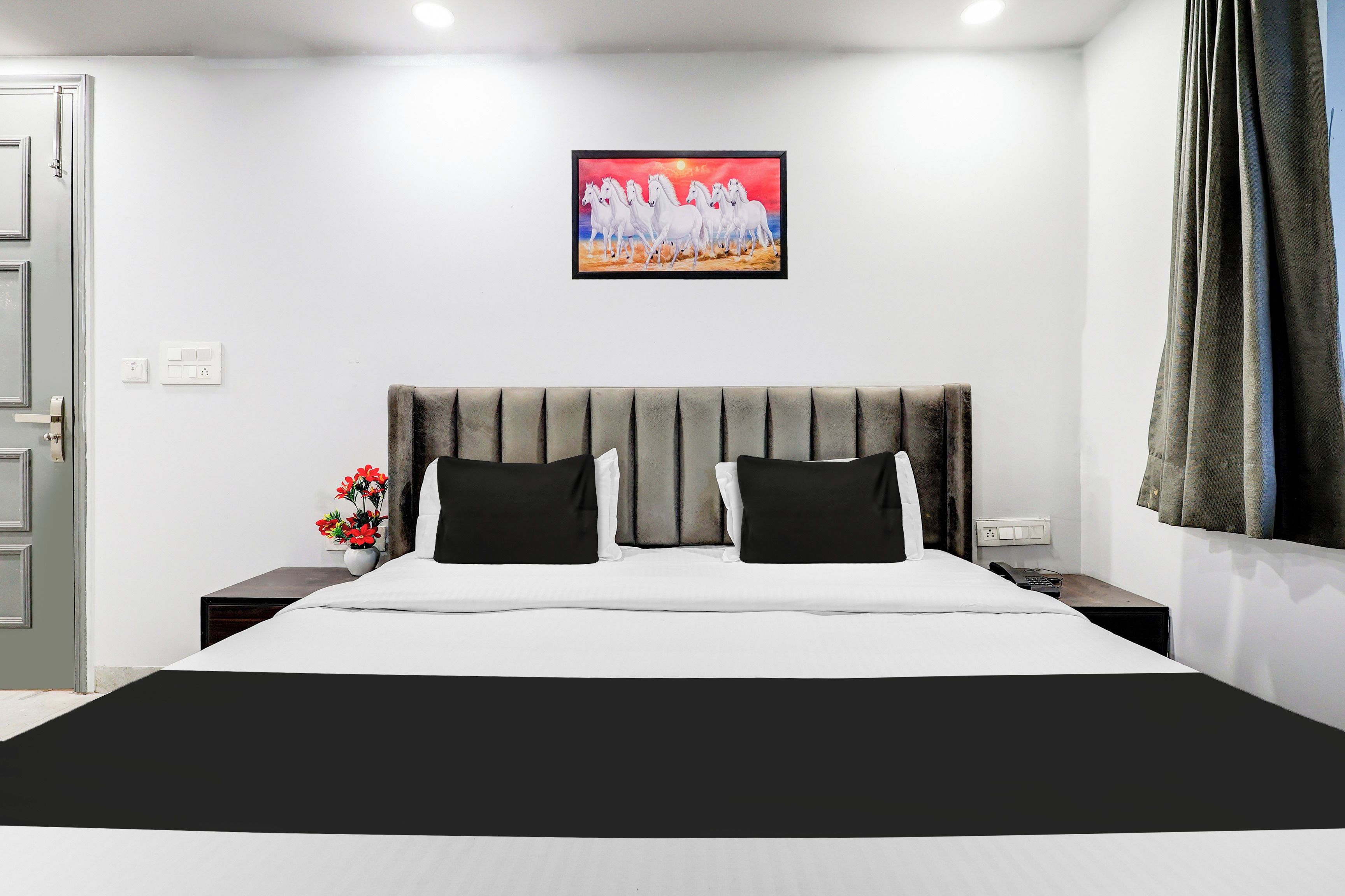 Una habitación de hotel con cama, mesita de noche, un florero con flores, un cuadro en la pared y una ventana con cortinas.