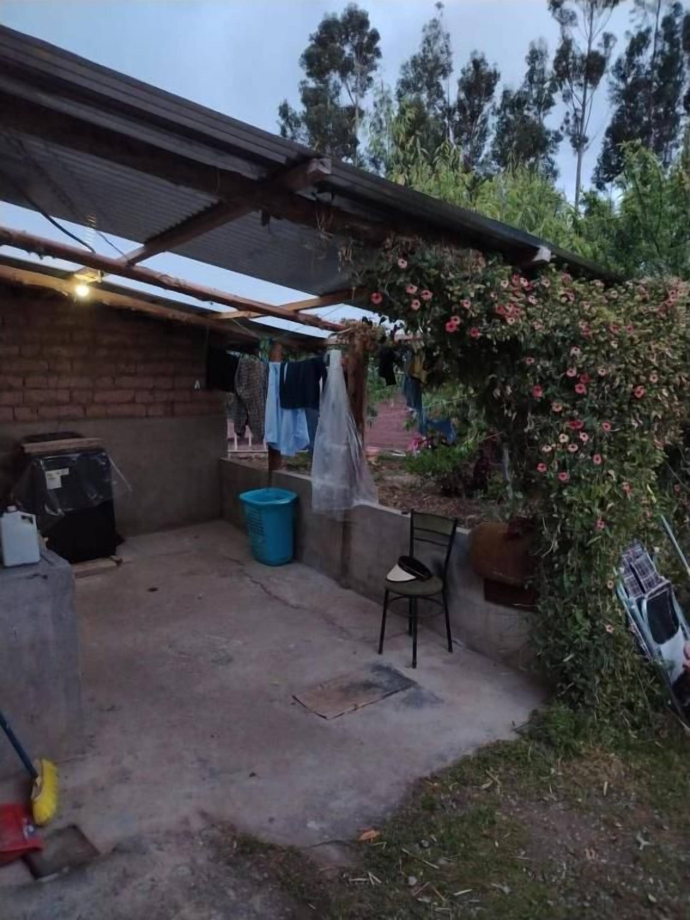 Área externa coberta com parede de tijolos, uma lixeira de plástico azul e uma cadeira.