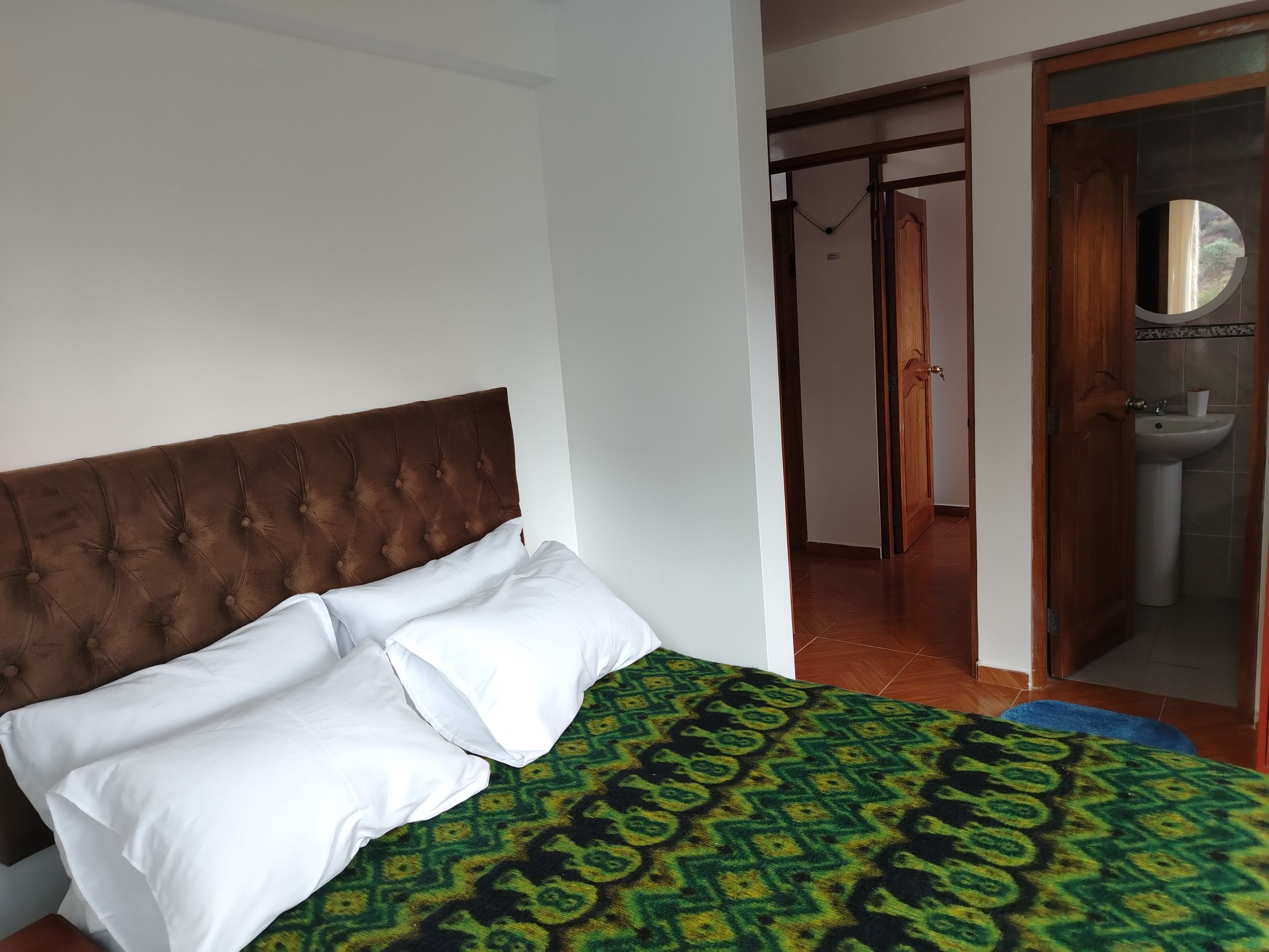 Un dormitorio con cabecera capitonada, una cama con ropa de cama blanca y verde estampada, y un baño visible a través de una puerta abierta.