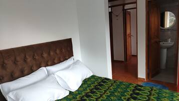 Un dormitorio con cabecera capitonada, una cama con ropa de cama blanca y verde estampada, y un baño visible a través de una puerta abierta.
