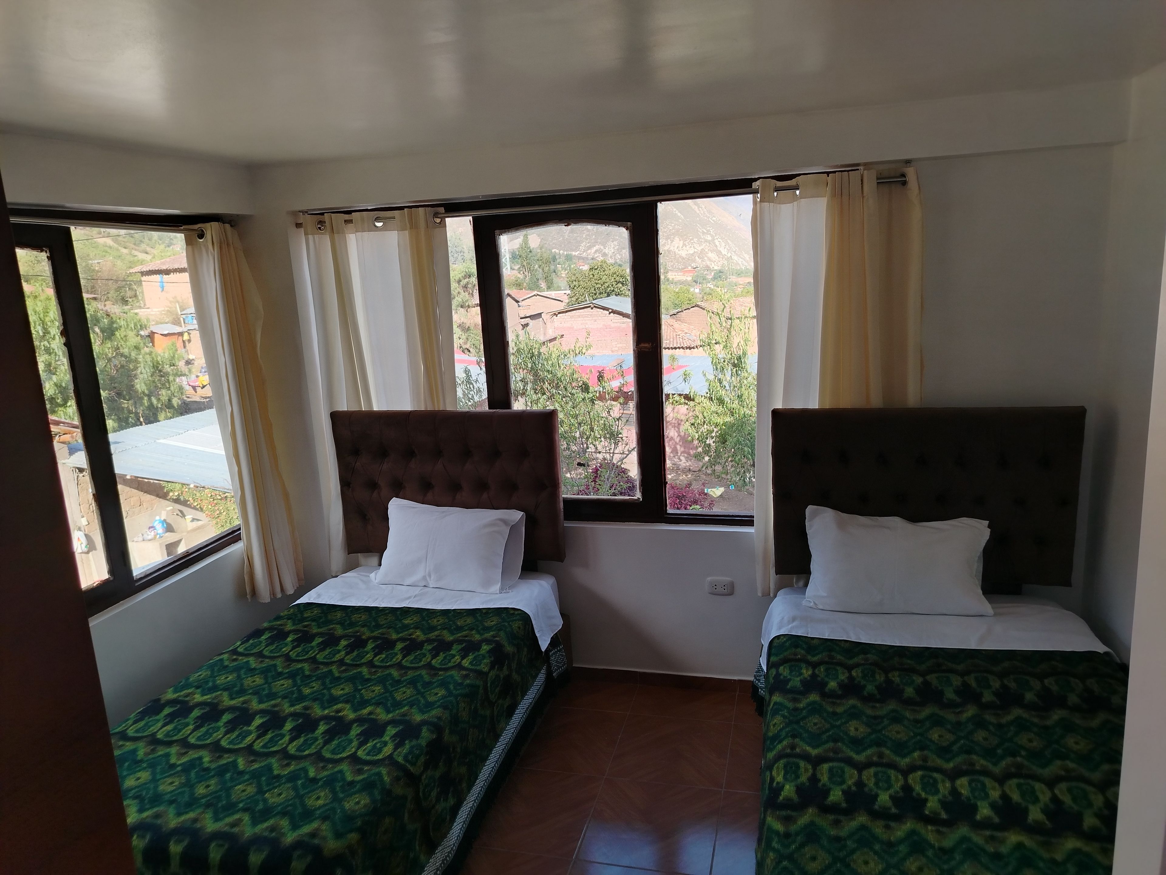 A room with two beds, each with a patterned bedspread, a window with curtains, and a view of a residential area outside.