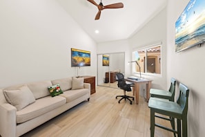 Ein Zimmer mit Deckenventilator, Schreibtisch mit Stuhl, einer Couch mit Kissen und Kunstwerken an den Wänden.