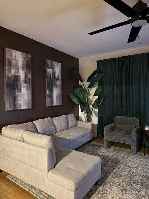 A living room with a sectional sofa, a small armchair, a large potted plant, and abstract wall art.