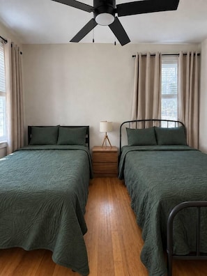 A room with two single beds, a ceiling fan, a bedside table with a lamp, and curtains on the windows.