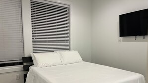 Een slaapkamer met een bed, witte beddengoed, een raam met jaloezieën, een aan de muur bevestigde televisie en een houten vloer.