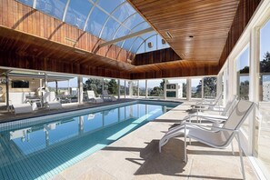 Une piscine intérieure moderne avec une eau d’un bleu cristallin, un plafond en bois et des chaises longues blanches.
