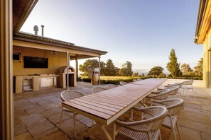 Une terrasse extérieure avec une longue table en bois, des chaises en osier, un barbecue et un téléviseur fixé au mur.