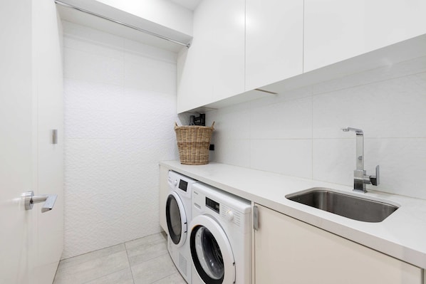 A compact laundry area with a washing machine, a sink, and a wicker basket on a countertop. - The Port Residence Luxury Port Melbourne Townhouse (Port Melbourne)