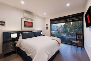A modern bedroom with a large bed, a nightstand, a chair, a TV, and a window with a view of the outside. - The Port Residence Luxury Port Melbourne Townhouse (Port Melbourne)