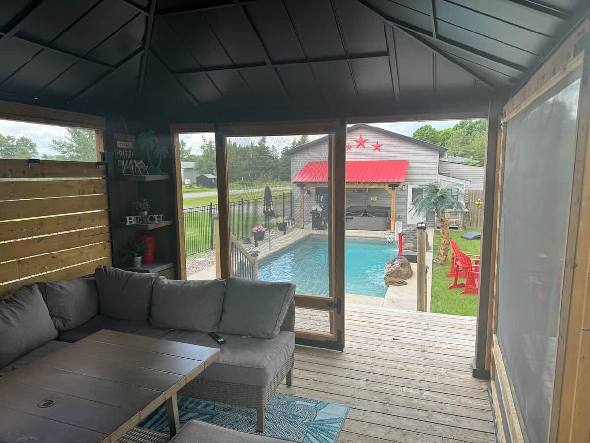 A covered outdoor seating area with a pool view, a patio, and a red-roofed building in the background.