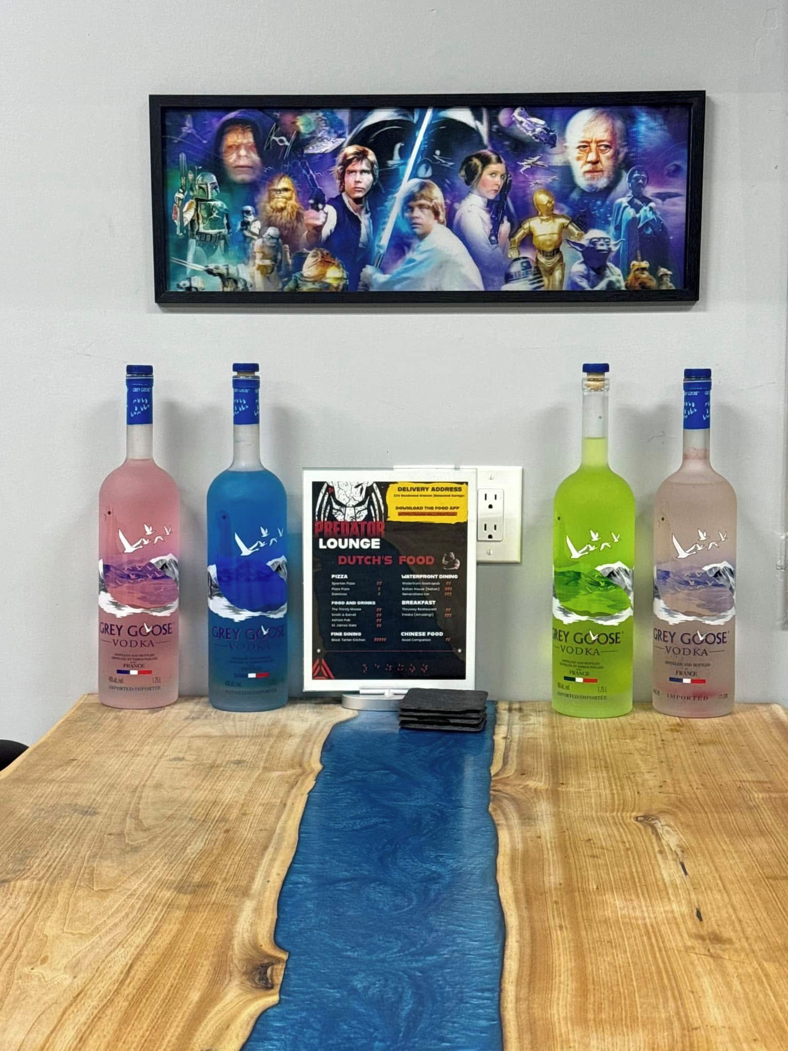 A room with a TV displaying a movie, a laptop, and four bottles of Grey Goose vodka on a wooden table.