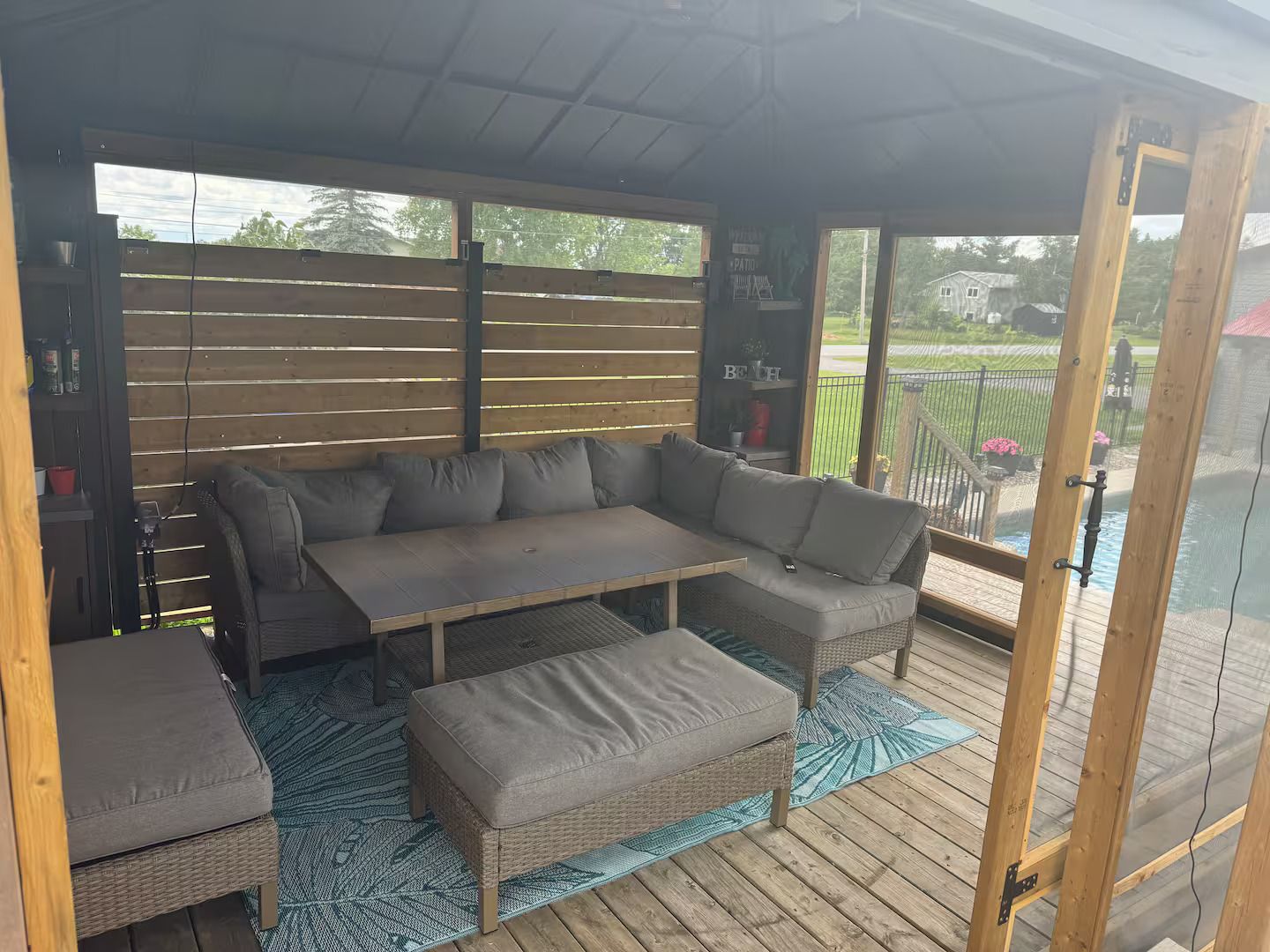 Un espace extérieur aménagé avec un coin salon couvert comprenant un canapé, un pouf et une table, une terrasse en bois et une vue sur un quartier résidentiel.