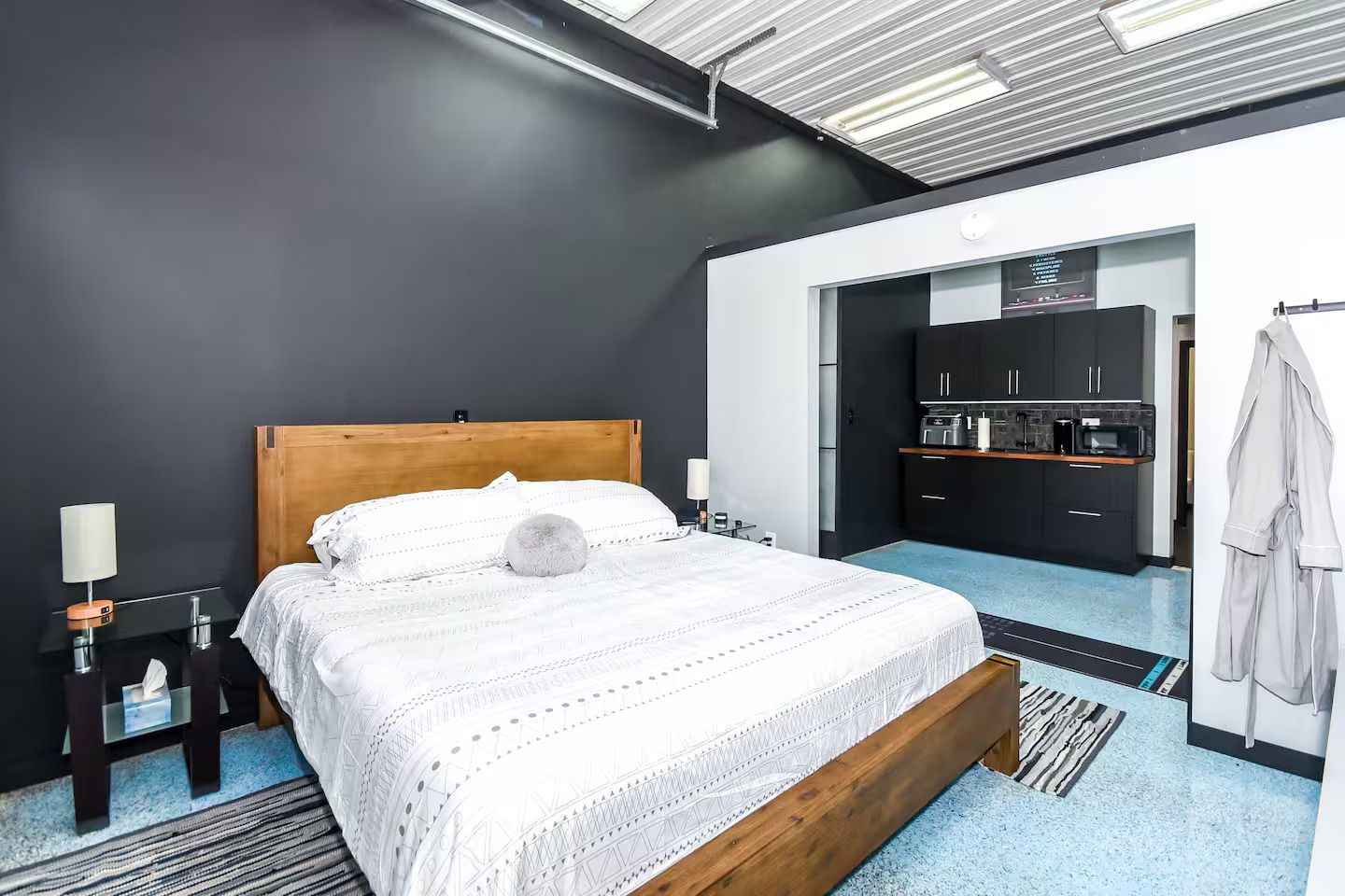 Habitación de hotel moderna con una cama de madera, una mesita de noche pequeña, una lámpara y vista al área de la cocina.