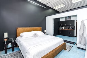 A modern hotel room with a wooden bed, a small bedside table, a nightstand, a lamp, and a view of the kitchen area.