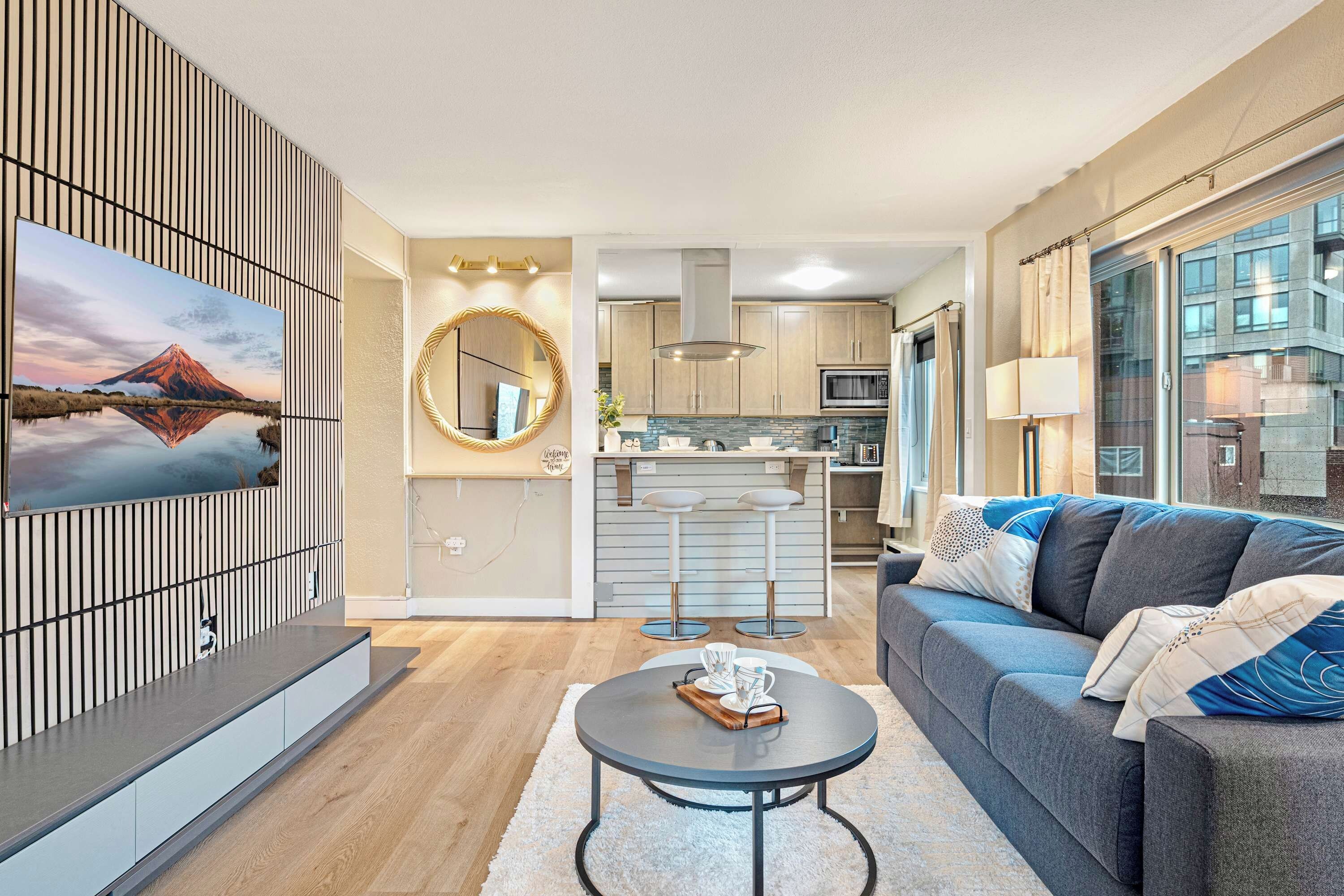 A modern living room with a sofa, a coffee table, and a TV mounted on the wall. There is a kitchen area with a bar and a dining table.