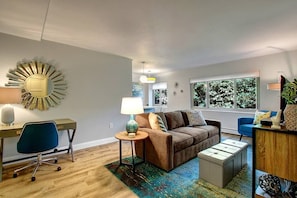 A modern living room with a brown sofa, a blue chair, a small round table with a lamp, and a large window with a view of trees. - Elektra (Seattle)