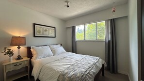 A bedroom with a bed, a nightstand, a lamp, a window with curtains, and a ceiling light fixture. - Bright & Cozy - The White Rock Gateway (Surrey)