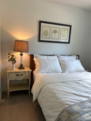 A neatly made bed with white bedding, a bedside lamp, a small bedside table with a vase of flowers, and a framed botanical print on the wall. - Bright & Cozy - The White Rock Gateway (Surrey)