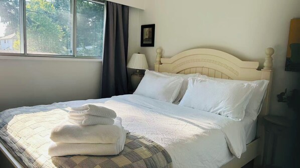 A neatly made bed with a headboard, a window with curtains, a bedside lamp, and a framed picture on the wall. - Bright & Cozy - The White Rock Gateway (Surrey)