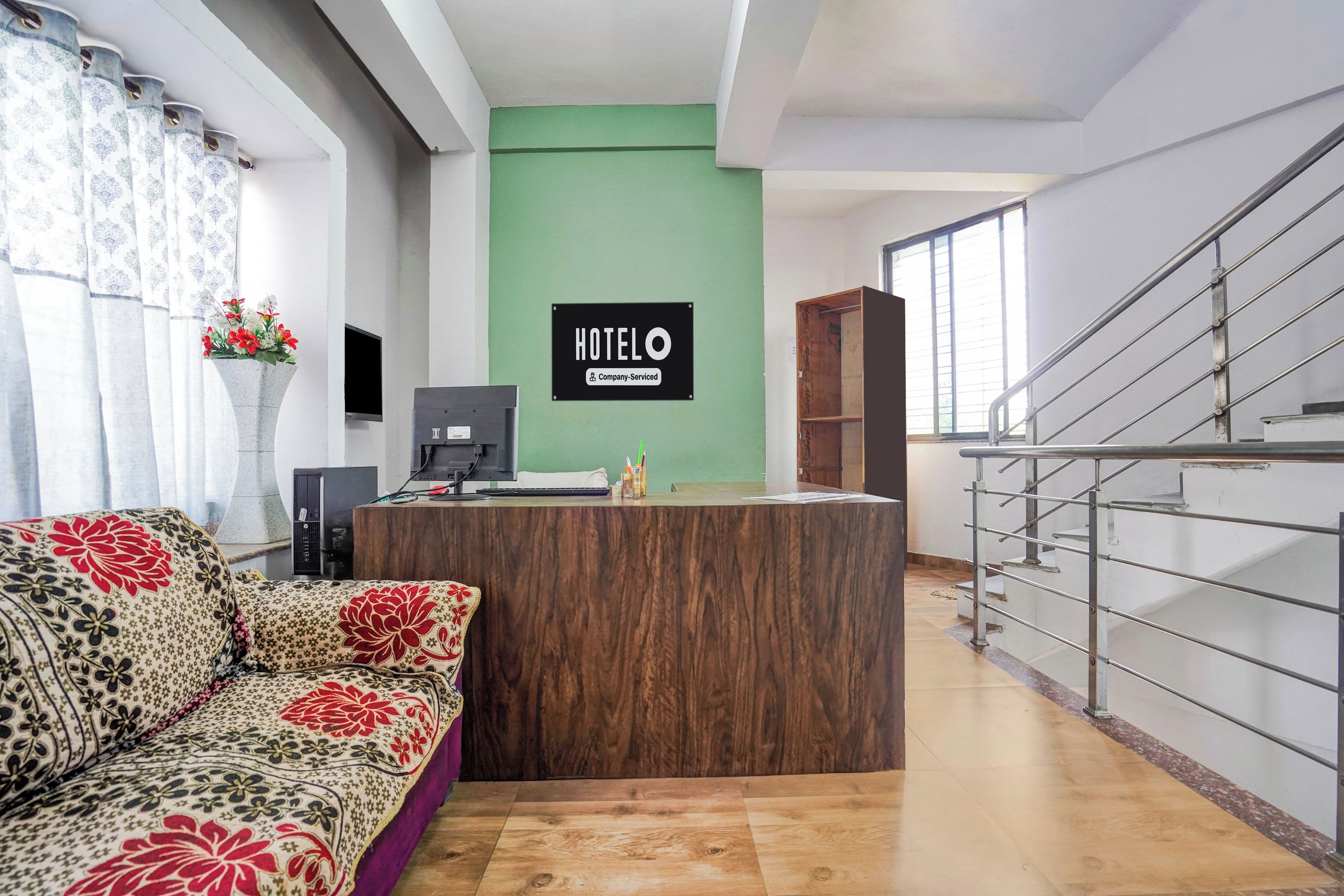 A hotel reception area with a wooden reception desk, a green wall, a staircase, and a sofa with floral upholstery.