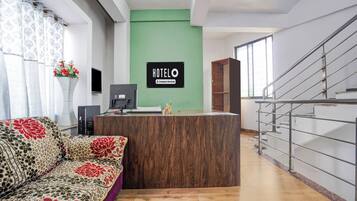 A hotel reception area with a wooden reception desk, a green wall, a staircase, and a sofa with floral upholstery.