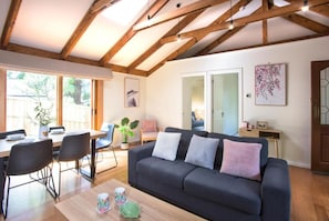 A modern living room with a dark sofa, a wooden dining table, and chairs. There is a large window providing natural light and a view of the outdoors. - Camellia Cottage (Olinda)