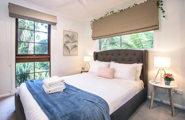 A bedroom with a large bed, two bedside lamps, a window with a view of greenery, and a wall-mounted artwork. - Camellia Cottage (Olinda)