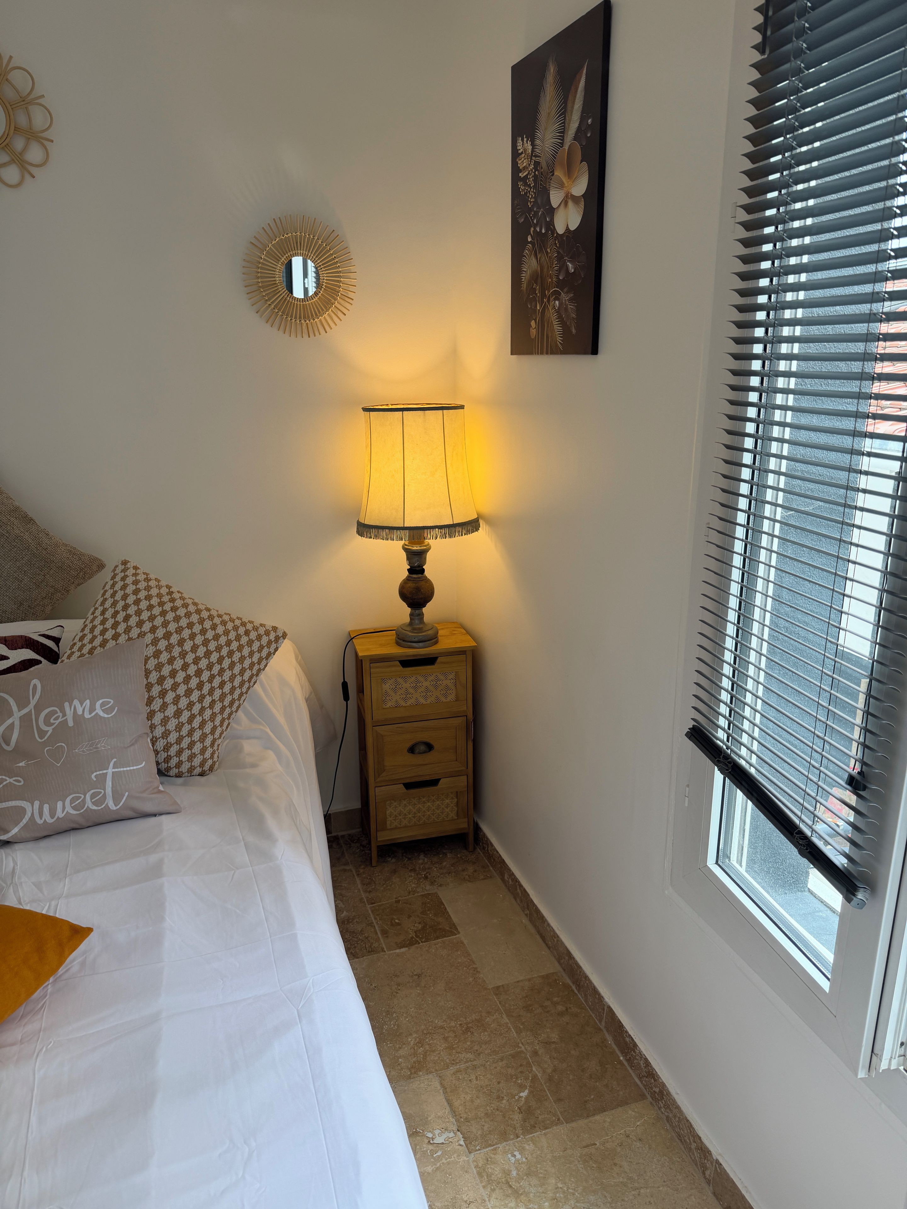Een slaapkamer met een bed, een nachtkastje met een lamp, een raam met zonwering en een decoratie aan de muur.