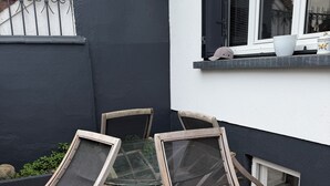 Une terrasse avec une table et des chaises, une fenêtre avec des volets et une petite marche menant à une porte.