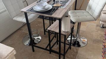 Zona pranzo con tavolo apparecchiato per quattro, una sedia imbottita bianca, angolo cottura con macchina del caffè e una finestra con persiane.