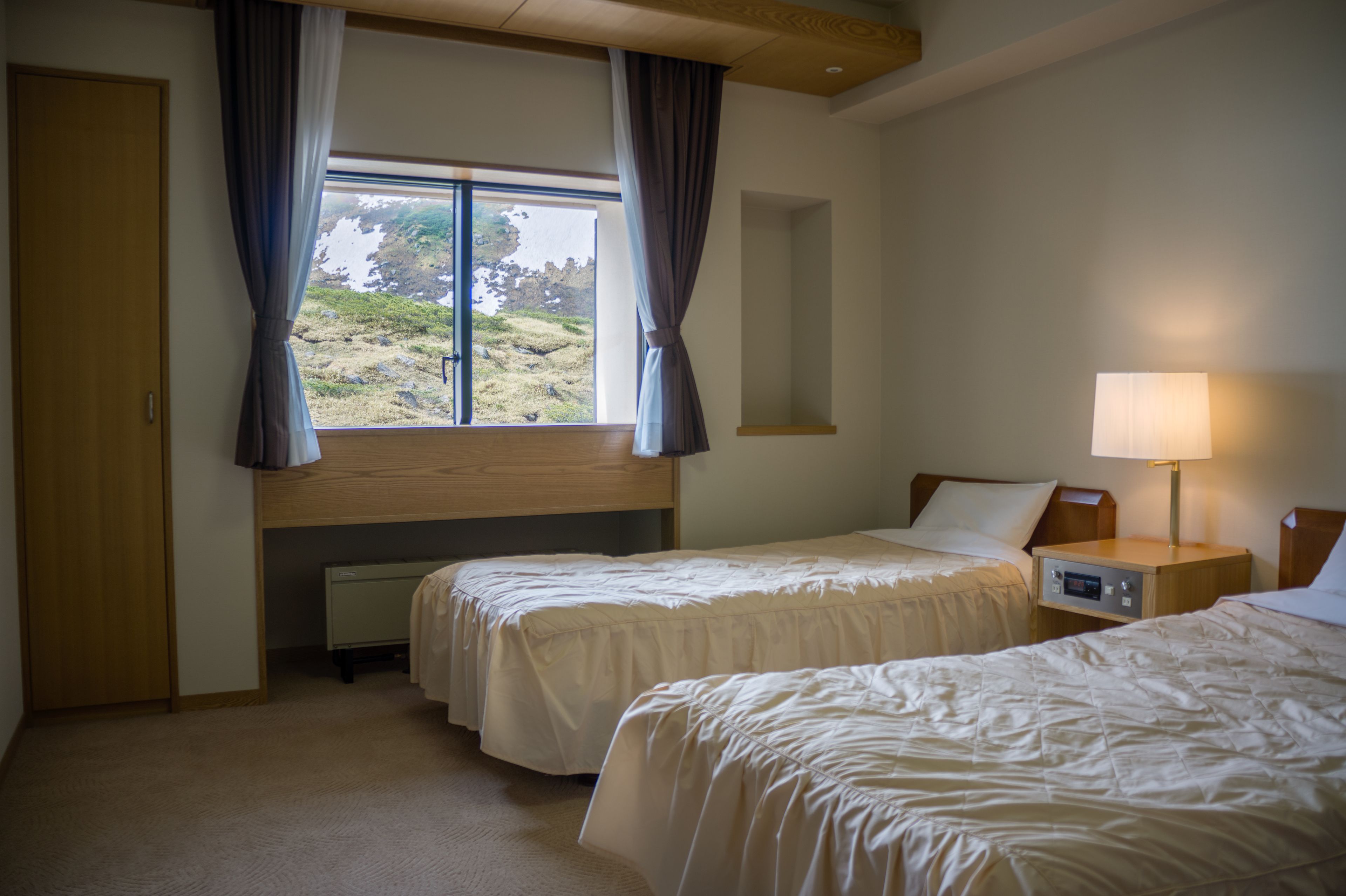 Une chambre d’hôtel avec deux lits, une fenêtre avec des rideaux, une table de chevet avec une lampe et un radiateur placé sous la fenêtre.