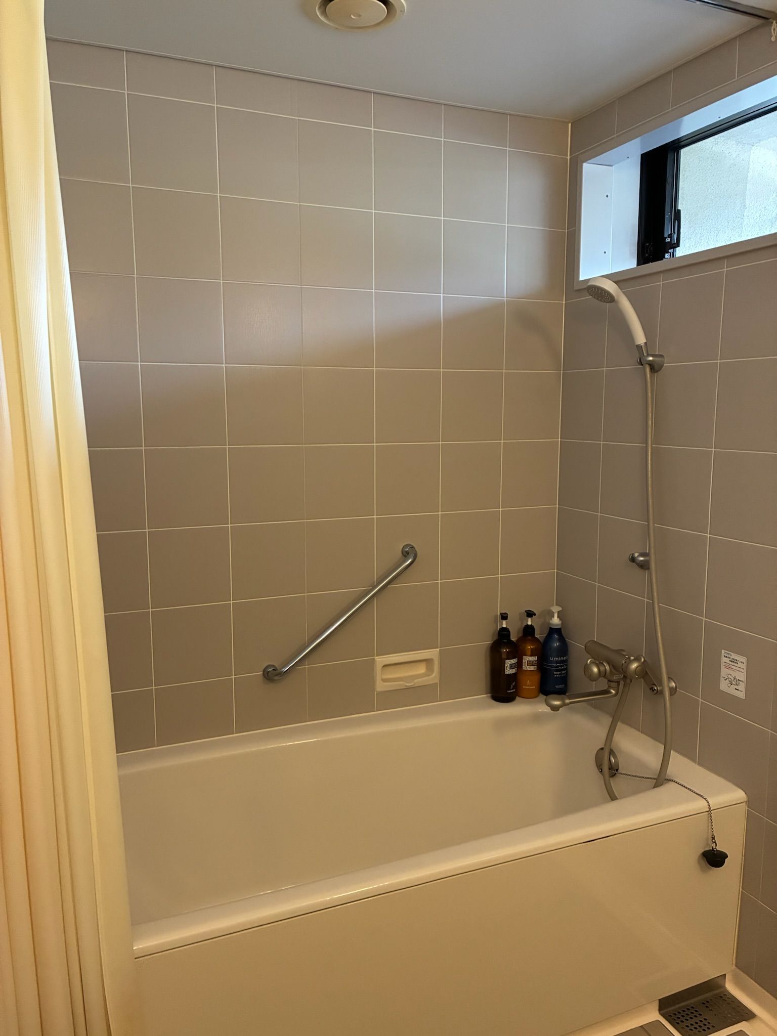 A bathroom with a bathtub, a shower, and a handrail. There are bottles of shampoo and conditioner on the wall.