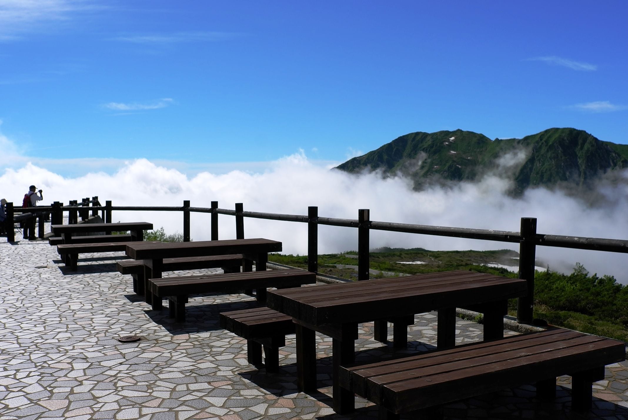 Bancos de madera al aire libre y mesas de picnic en un área empedrada con vistas a una montaña y nubes.