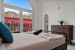 A bedroom with a bed, a dresser, a mirror, and a view of buildings through large windows. - Grand Galleria — Old San Juan Private Patio (San Juan)