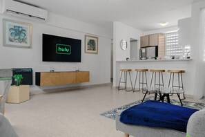 A modern living room with a flat-screen TV, a kitchen area with bar stools, and a dining table. - Private Patio 2BR Near Ocean Park Beach (San Juan)