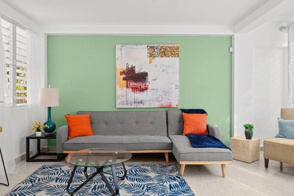 A modern living room with a grey sofa, orange and blue cushions, a glass coffee table, and a green accent wall. - Private Patio 2BR Near Ocean Park Beach (San Juan)