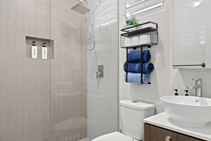 A modern bathroom with a glass shower enclosure, a white sink, and a towel rack with folded towels. - Private Patio 2BR Near Ocean Park Beach (San Juan)