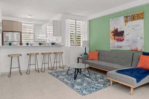 A modern living room with a grey sofa, a coffee table, and a zebra-patterned rug. There are four wooden stools and a kitchen area with a refrigerator and a window with blinds. - Private Patio 2BR Near Ocean Park Beach (San Juan)