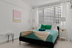 A bedroom with a bed, two bedside tables, a lamp, a window with blinds, and a wall-mounted artwork. - Private Patio 2BR Near Ocean Park Beach (San Juan)