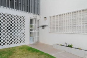 Ein Gebäude mit weißer Fassade, einer nummerierten Tür, einem Fenster mit Gitterstäben und einem kleinen Garten.