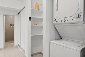 Ein Waschraum mit einer Frontlader-Waschmaschine, einem Regal mit Dekoration und einer Tür zu einem Badezimmer.