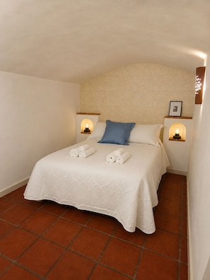 A neatly made bed with a white bedspread and two pillows, one blue and one white, in a room with terracotta floor tiles and a white ceiling.